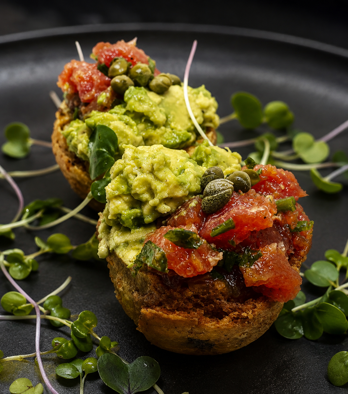 Crostini topped with avocado, citrus segments and capers arranged on a dark plate with micro herbs