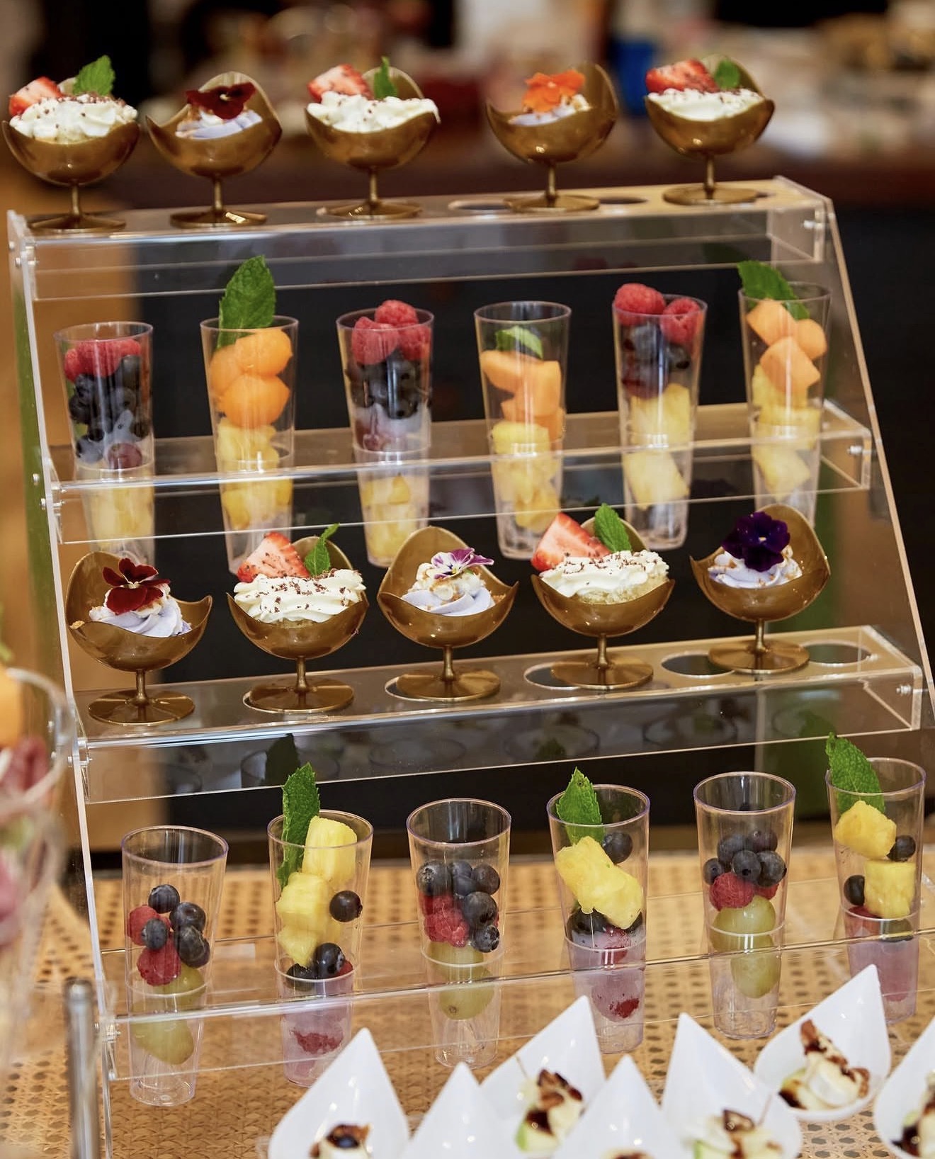 An elegant tiered display of individual desserts in gold cups and fresh fruit shooters