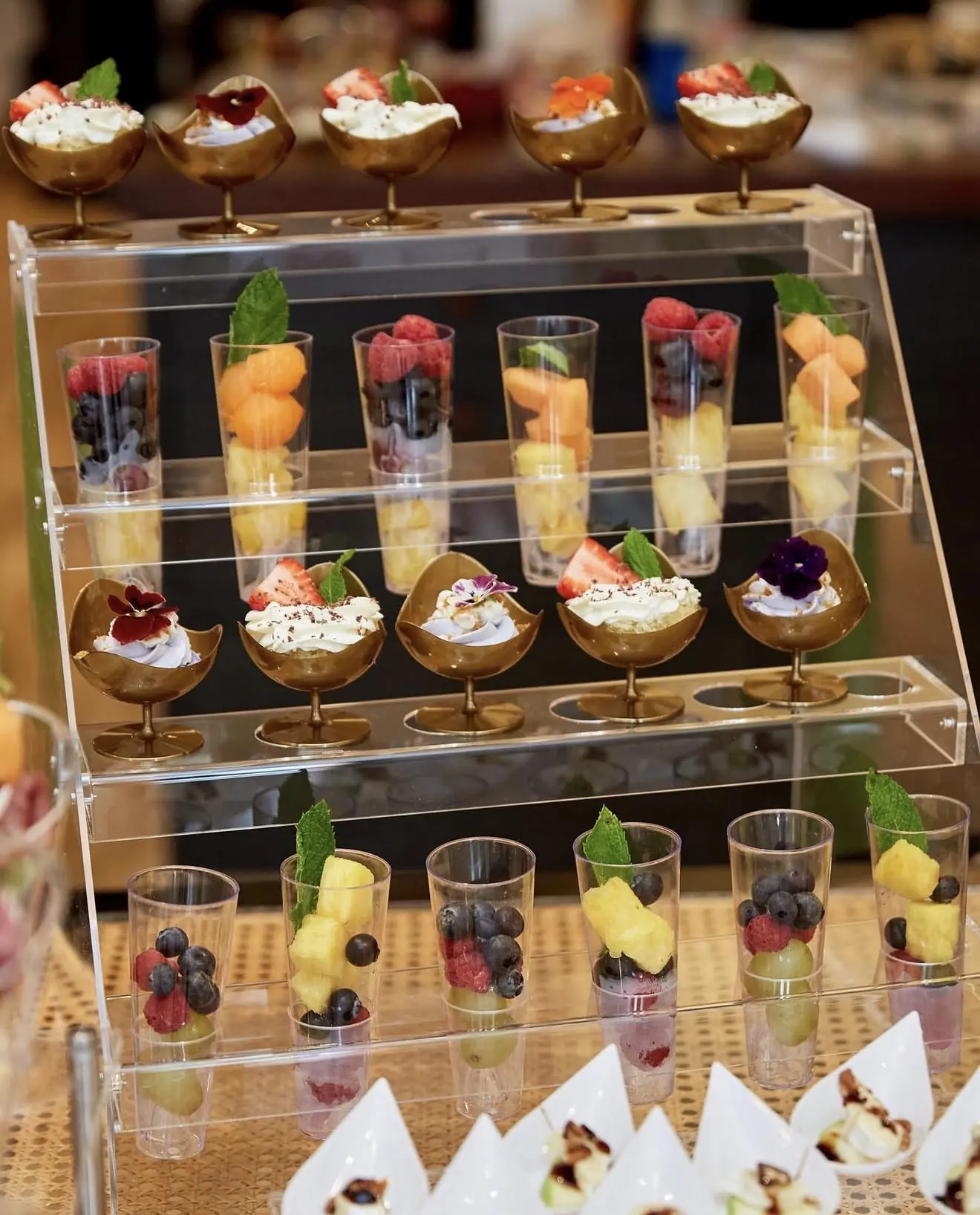 An elegant tiered display of individual desserts in gold cups and fresh fruit shooters